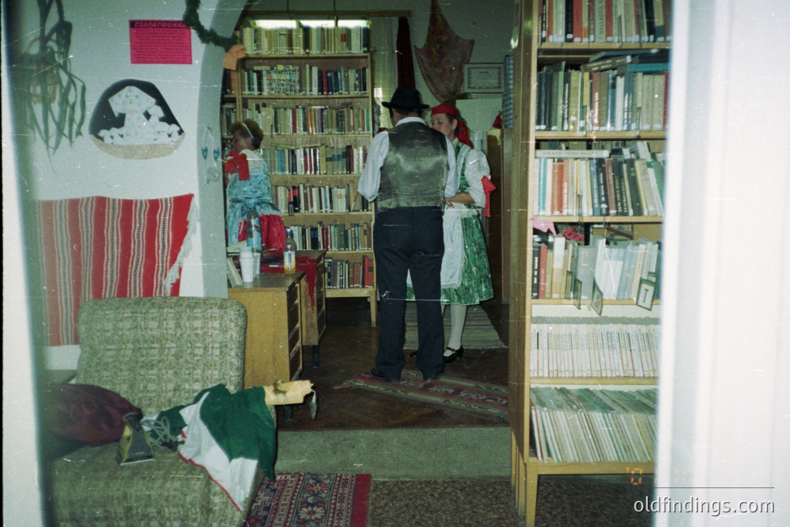 A man in a dark suit and vest stands facing a woman in traditional folk dress in a room filled with bookshelves. The woman wears a green dress, red sash, and matching hat. A patterned rug and armchair are visible in the foreground. Likely a cultural performance or demonstration.