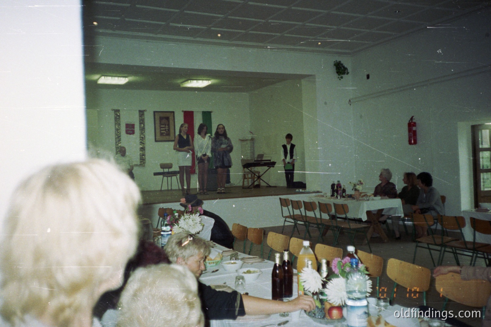 Indoor scene of a gathering, likely a community event or performance. A small stage features three figures; two women and a boy in a suit. Tables with white cloths and floral centerpieces fill the room. Appears to be 1970s-80s based on clothing and photographic style.