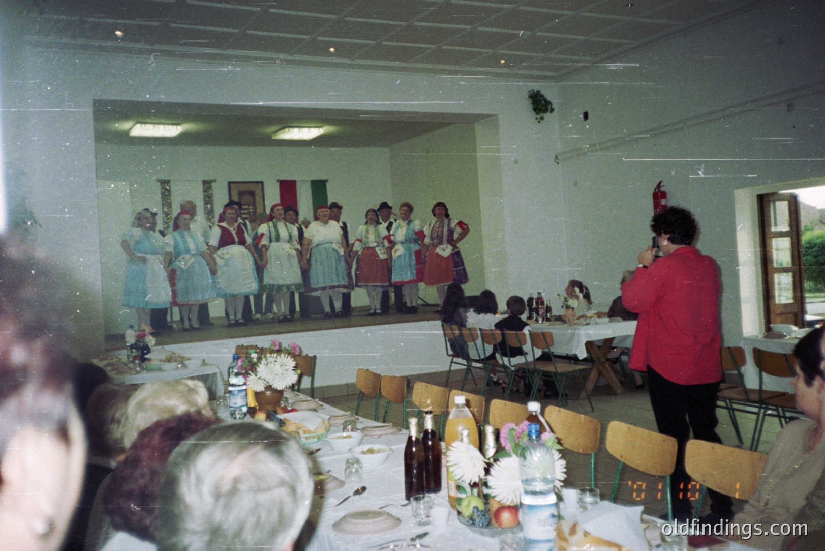 A group performance on stage featuring individuals in traditional folk attire. Seated audience observes from tables laden with food and drink. Likely a cultural celebration; image reflects 1970s or 80s photography style. Venue appears to be a community hall.