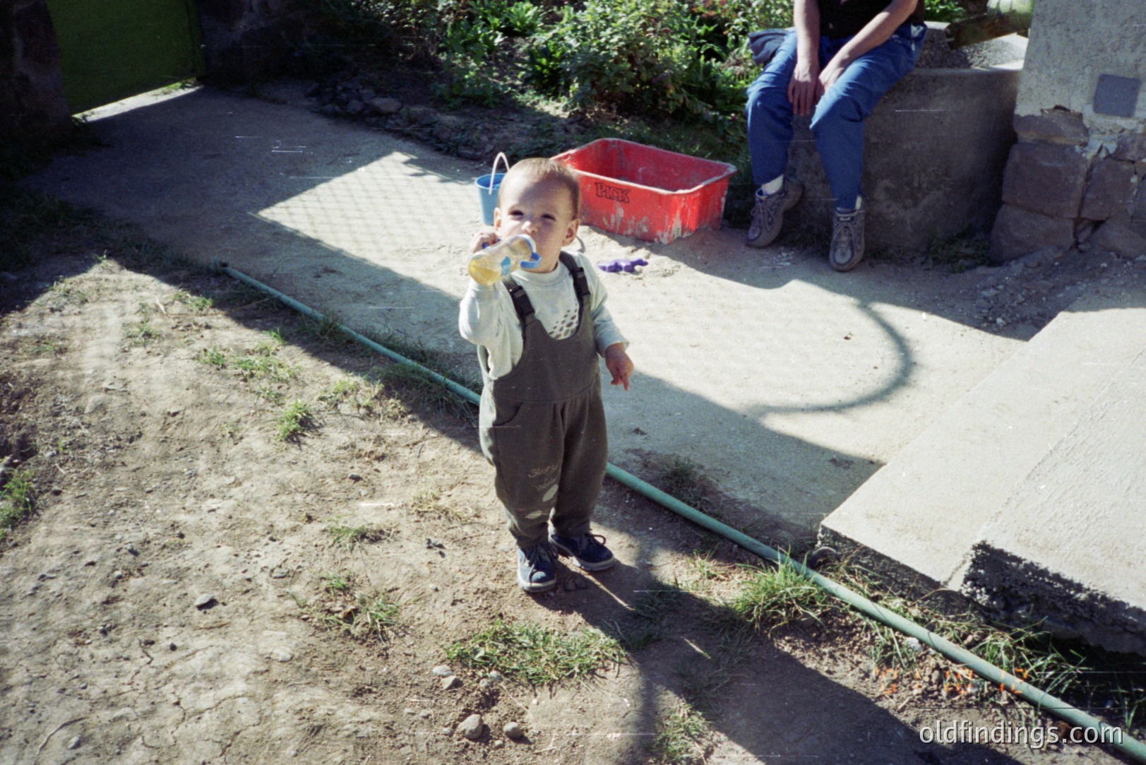 A young child, wearing overalls and dark shoes, stands holding a blue bucket. A seated individual in jeans and work boots is visible in the background. The scene appears to be a sunlit, unpaved area. Likely a home or rural setting.