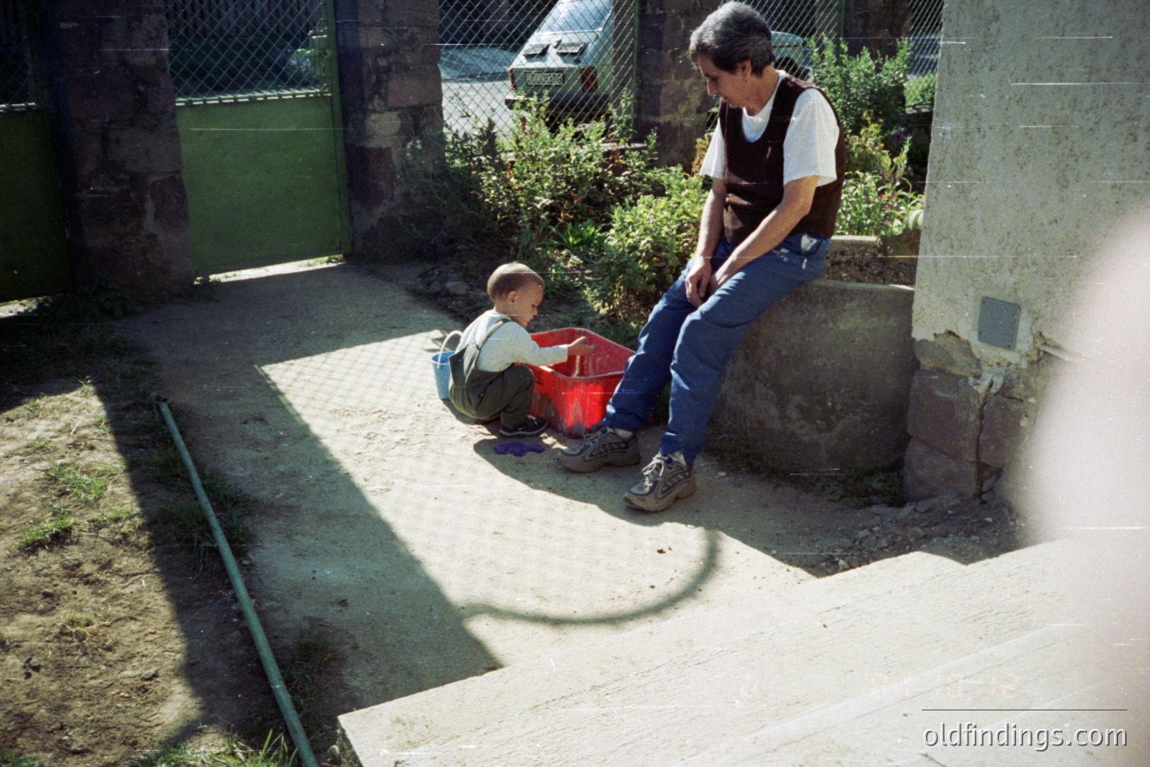 A young boy in a red shirt & shorts sits beside a man wearing jeans & a vest, both outdoors. They occupy a concrete courtyard shaded by a building. The scene appears residential, with greenery visible. Appears to be a candid moment, perhaps 1980s.