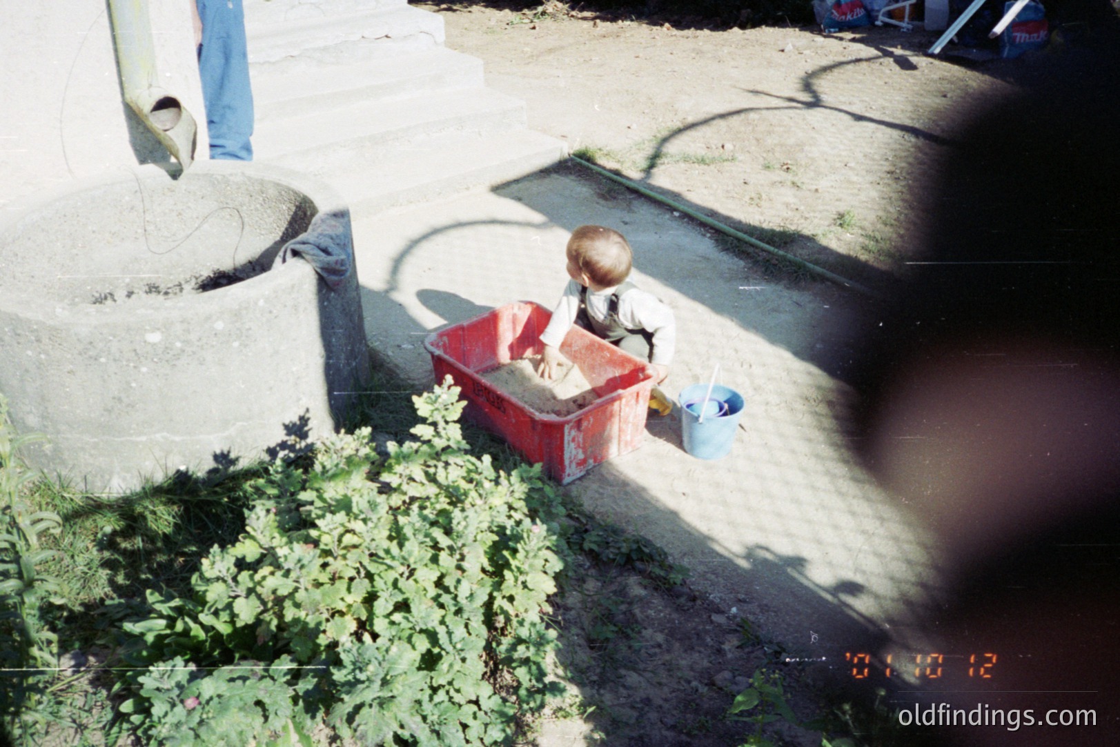 A young boy sits in a red plastic bin, playing with sand near a concrete structure (possibly a well or fountain) and a low stone wall. A blue bucket sits beside him. Appears to be an outdoor domestic scene. Timestamp visible in the lower right corner: "08/08/12." Likely 2000s.