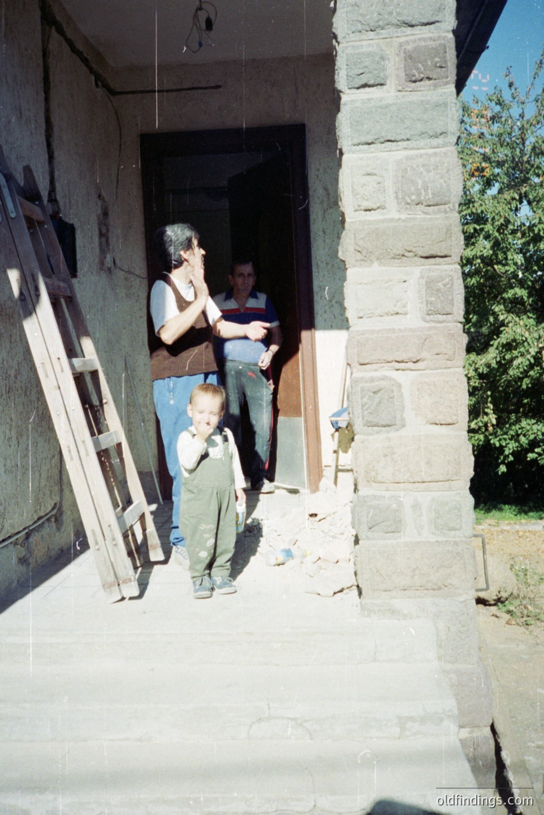 A young boy in overalls stands on a stone porch. Two men stand inside an open doorway, seemingly observing construction work. Weathered stone facade and a ladder indicate ongoing building or renovation. Likely a residential scene, potentially in Eastern Europe. Appears to be a candid moment, possibly 1970s-1980s.