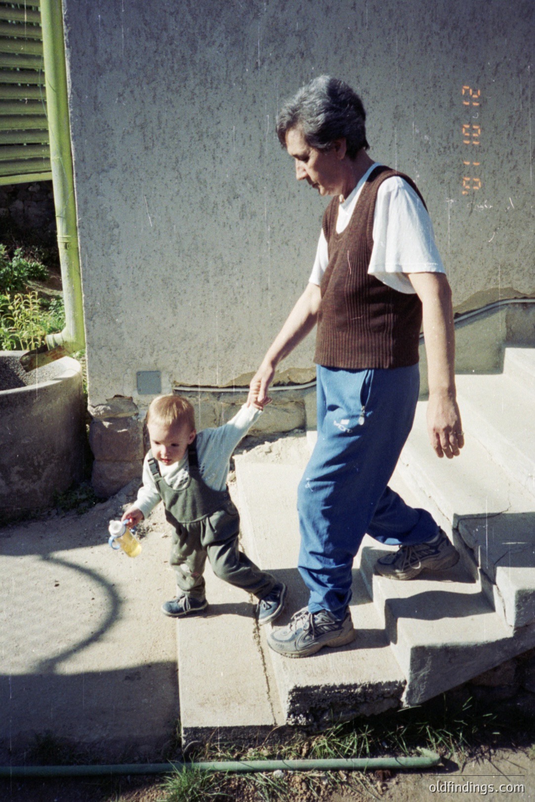 A woman assists a toddler descending concrete steps. The child, in overalls and a hat, carries a bottle. The woman wears a sweater vest over a white shirt and jeans. Stairwell facade suggests mid-century architecture. Date visible: 8/10/’83. Candid family moment.