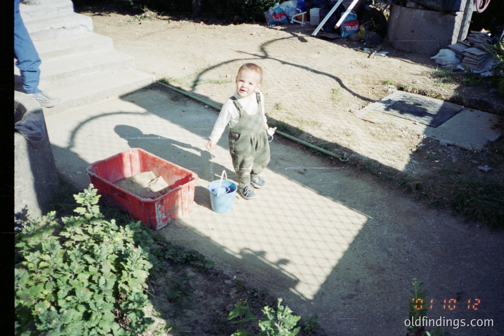 Young boy in overalls stands in a sun-drenched yard, alongside a red plastic tub and a blue bucket. Appears to be a construction site or renovation project underway. Likely 1990s, possibly Eastern European setting given the photographic style and setting.