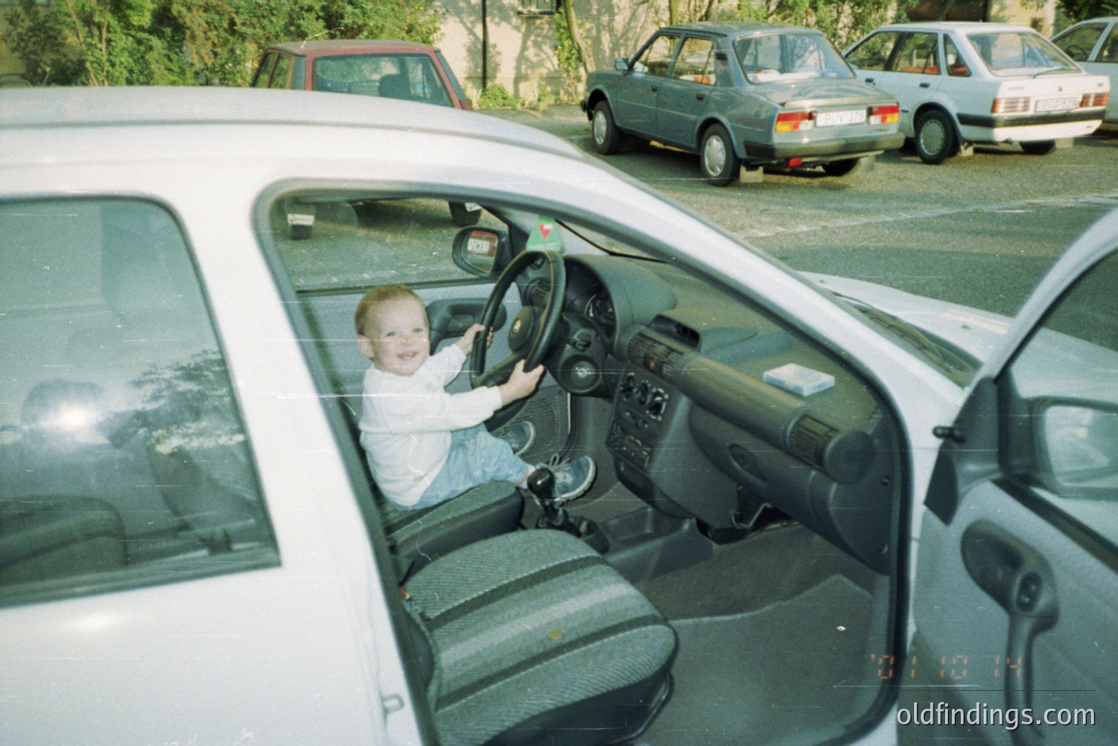 A young child, appearing to be a toddler, sits in the driver’s seat of a white sedan, mimicking driving with both hands on the steering wheel. The interior shows a 1990s car design. Other vehicles are visible outside. Likely a candid snapshot.