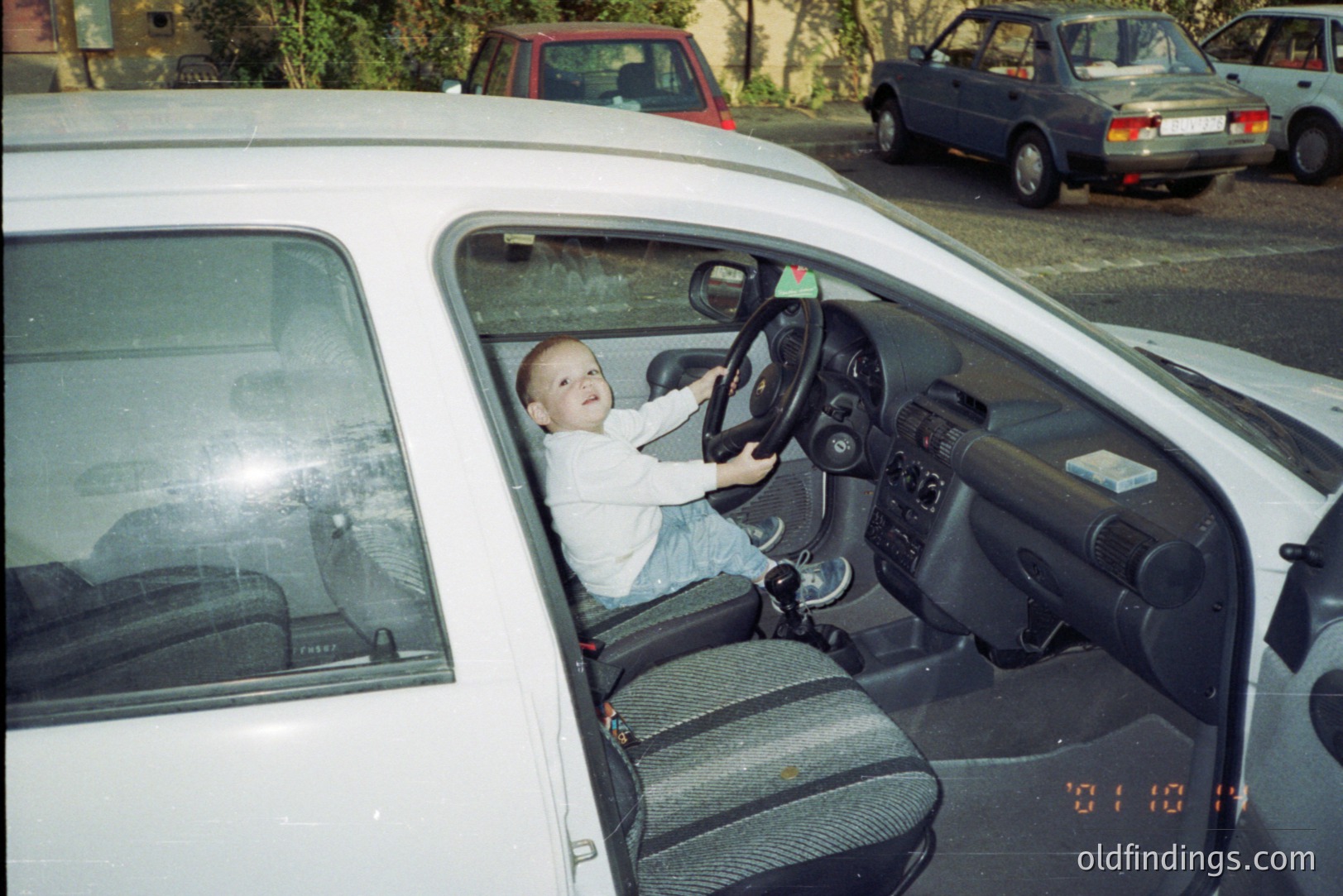 A young boy sits in the driver's seat of a white Volkswagen, playfully gripping the steering wheel. Visible date stamp indicates photo was taken 8/11/1998. Interior shows typical 1990s automotive design. Two cars are visible in the background. Nostalgic snapshot of childhood.
