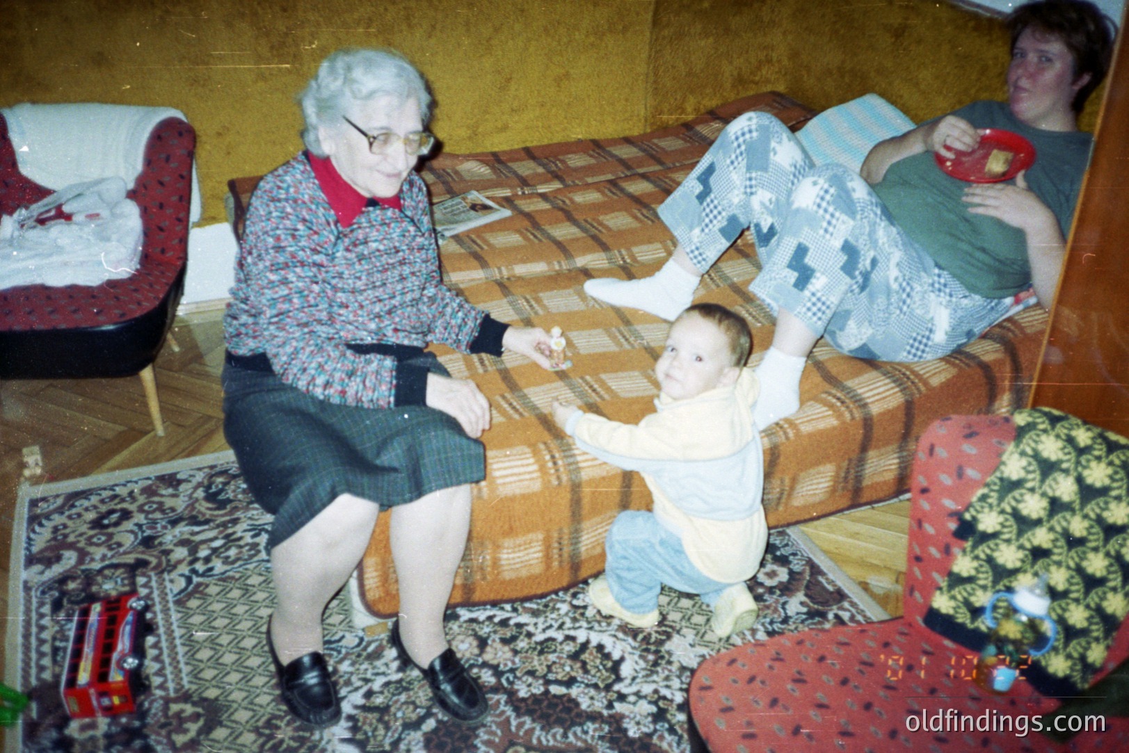 An elderly woman in a patterned dress and black shoes sits with a toddler reaching towards her. A young woman in casual attire reclines on a patterned sofa. Likely a domestic interior, possibly 1970s-1980s, based on decor and clothing styles. Family portrait with a candid, informal feel.