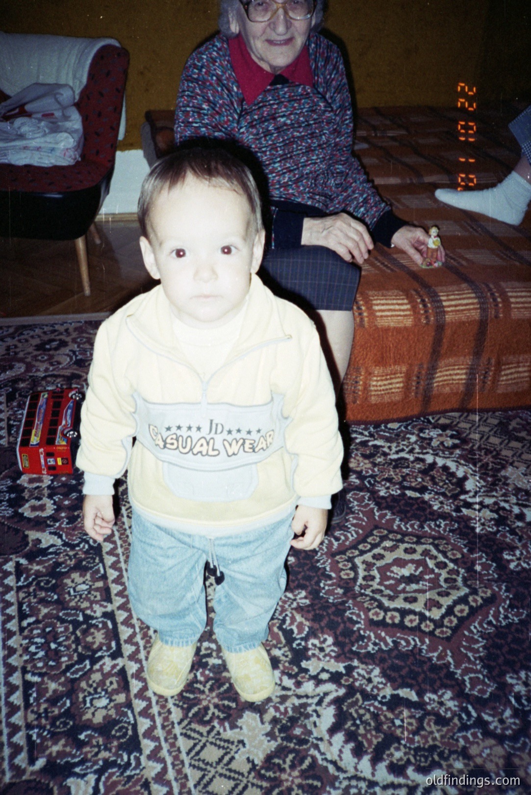 Infant stands facing the camera in casual wear: pale yellow sweatshirt & faded blue jeans. A patterned rug fills the foreground. An elderly woman is visible in the background, holding a small toy. Likely a family snapshot from the 1990s.