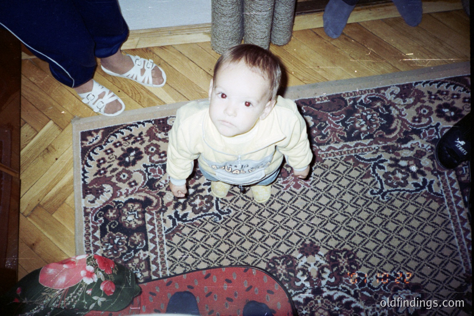 Infant wearing a cream-colored sweater stands on a patterned area rug with floral and geometric elements. The photograph’s perspective is from above. Visible are adult feet in sneakers and a partially visible dark shoe. Likely a candid, personal snapshot.
