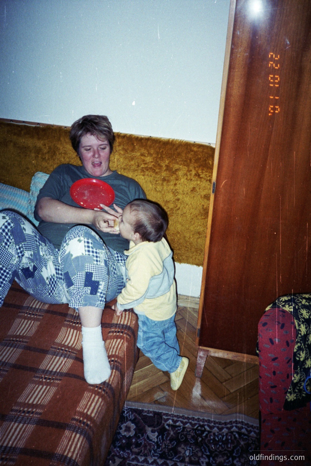 A young boy in overalls playfully reaches toward a woman seated on a patterned sofa. She's wearing a patterned pajama top and socks, holding a blue plate. The room appears to be a domestic interior, likely the 1980s. The image captures a candid, family moment.