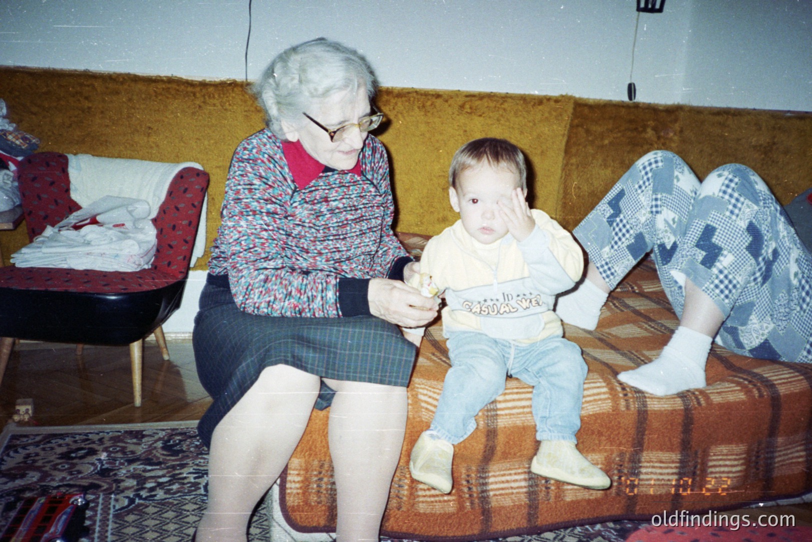 An elderly woman with a red sweater and plaid skirt sits on a floral sofa, holding an object in front of a seated toddler. The toddler wears a "Casual Sport" sweatshirt & tan pants. Likely a family snapshot, circa 1980s, showcasing domestic life. Interior features vintage furniture and decor.
