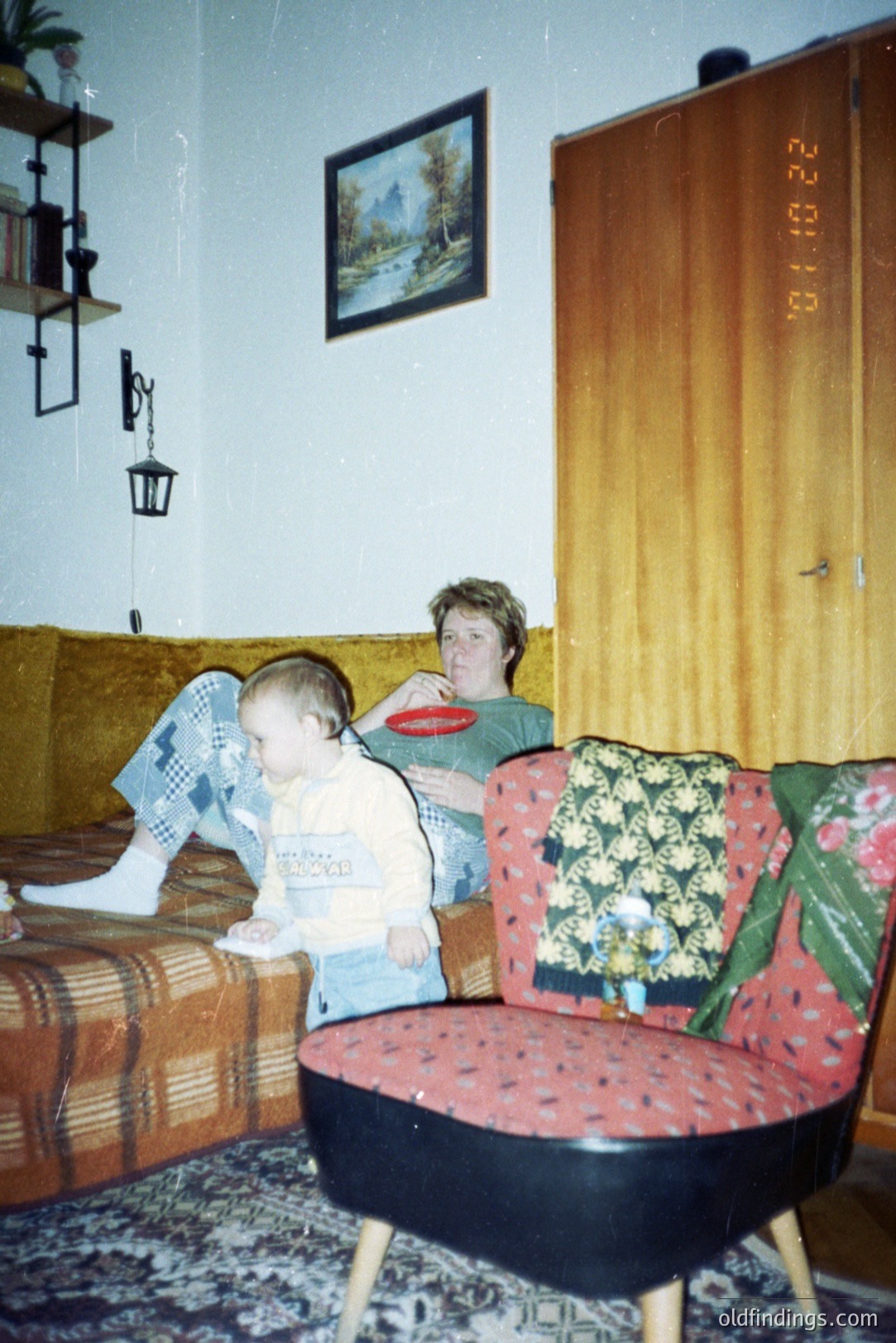 A young boy in denim overalls sits on a patterned sofa next to a woman holding a plate. The room features dated decor: a landscape painting, shag carpeting, and a distinctive round chair. Appears to be an interior shot from the 1970s. Typical family snapshot style.