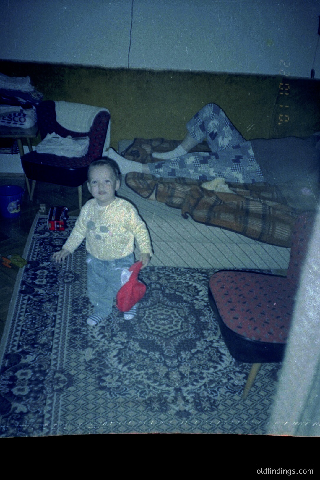 A young toddler stands on a patterned rug, holding a red toy. A woman is partially visible on a couch in the background, displaying a 1970s style interior. Likely a snapshot, the image exhibits characteristic color cast.