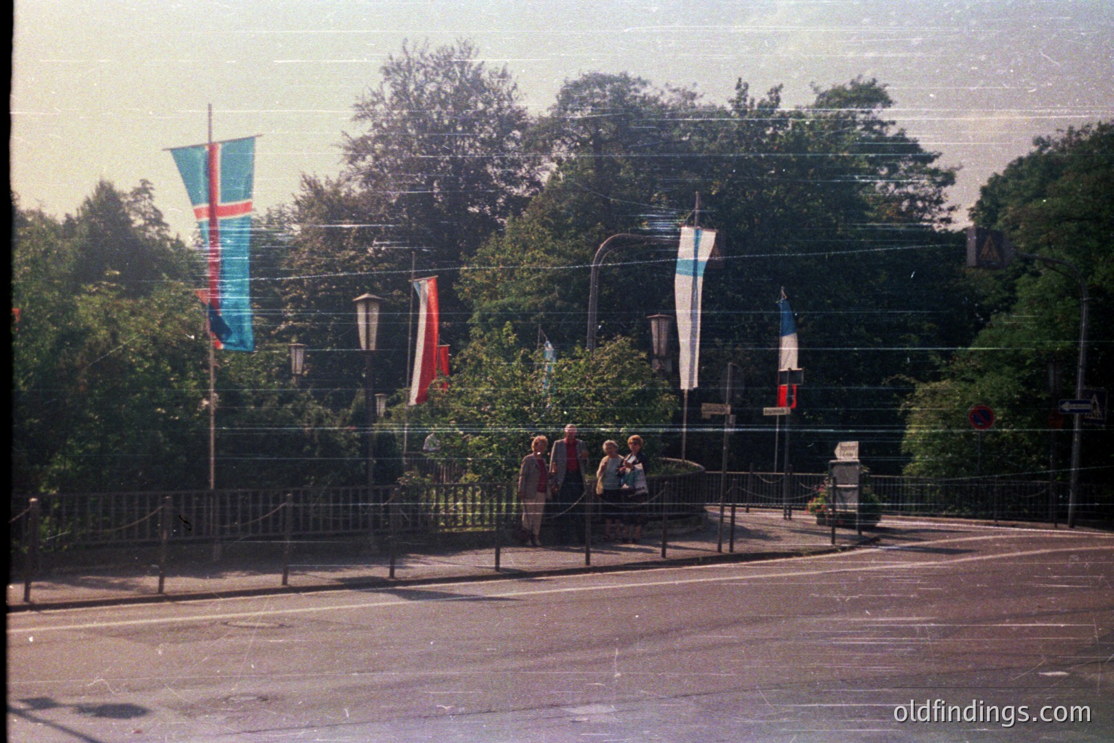 A group of people walk along a paved pedestrian walkway with metal railing, lined with vibrant flag banners. Lush greenery fills the backdrop behind the flags. Appears to be an elevated overlook or bridge. Likely a tourist area. Film grain suggests a 1970s origin.