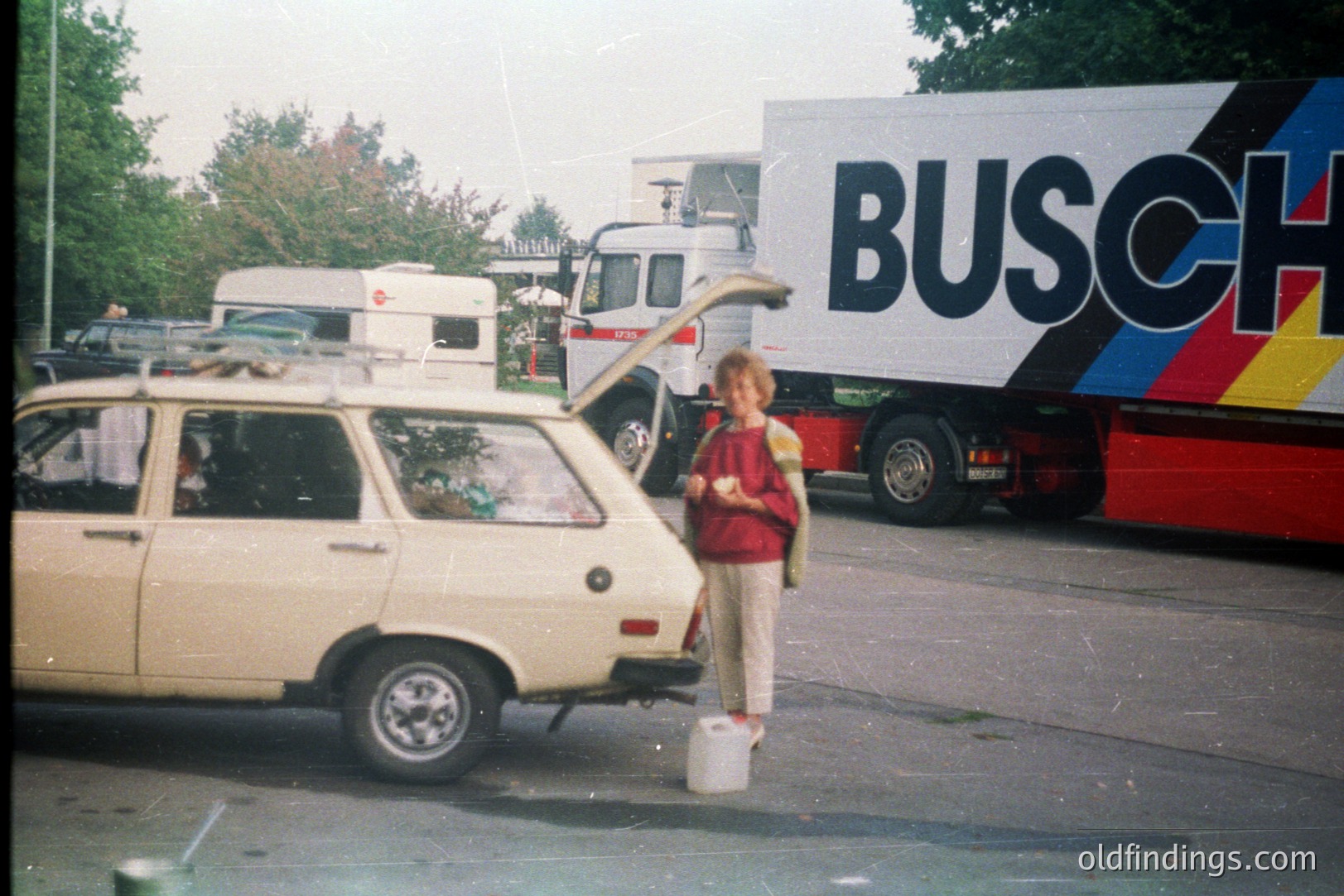 A young boy stands beside a beige Lada estate car, a bucket at his feet. A large "BUSCH" branded truck is visible in the background, suggesting a transportation or logistics context. The car has a roof rack with luggage. Appears to be an outdoor, possibly roadside, location. Likely 1970s or 80s.