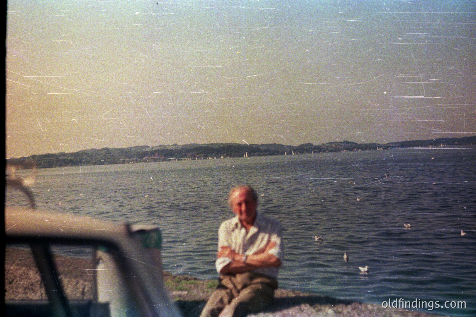A seated older man, arms crossed, overlooks a wide body of water with distant hills. The photograph exhibits characteristics of faded color film, likely 1960s-1970s, showing light scratches & dust. A car's side mirror is visible in the lower left corner. Scenic view with potential coastal or lakeside setting.