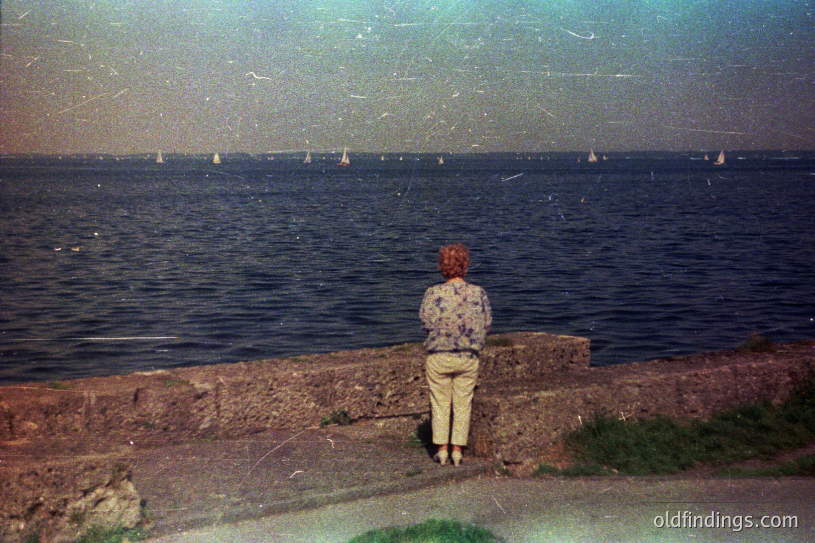 A figure with auburn hair stands overlooking a wide expanse of ocean, observing numerous sailboats on the water. The scene features a textured stone retaining wall with sparse grass and a paved walkway. Image exhibits signs of age/wear, with visible film grain and scratches. Likely a vacation snapshot, 1970s-1980s.