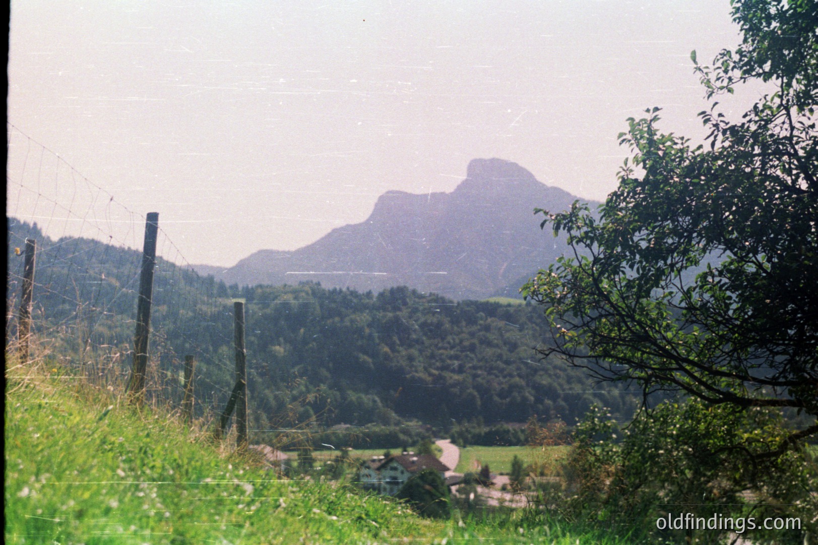 Hilly, forested landscape with a distinctive, jagged mountain peak dominating the view. A small village and reservoir are nestled amongst the greenery. Appears to be a vintage film photo, likely from the 1970s or 80s. A fence and foliage frame the scene.