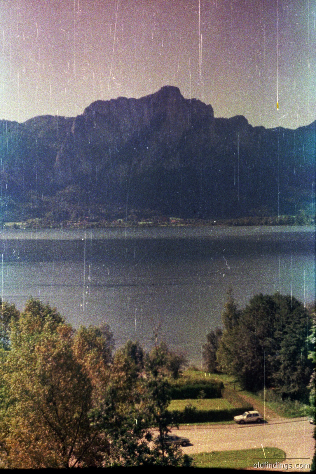 Lush foliage frames a scenic view of a lake reflecting imposing mountains. A vintage car is parked on a paved drive leading toward the water's edge. Likely taken in the 1960s or 70s, it evokes a sense of mid-century travel & leisure.