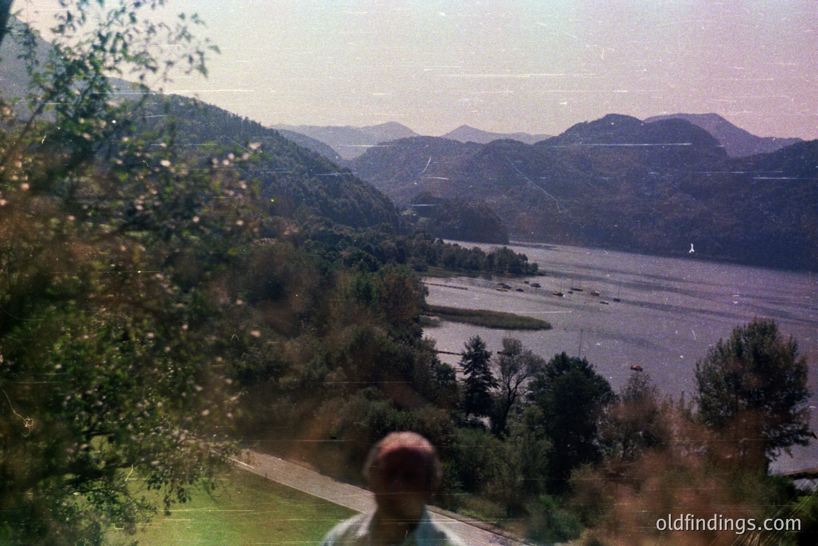 Scenic view of a river valley, likely the Hudson River, framed by lush, forested hills. A person stands at the edge of a lawn, appearing to observe the landscape. Photo exhibits signs of age with dust and color shift. Suggestive of travel photography, circa 1970s.