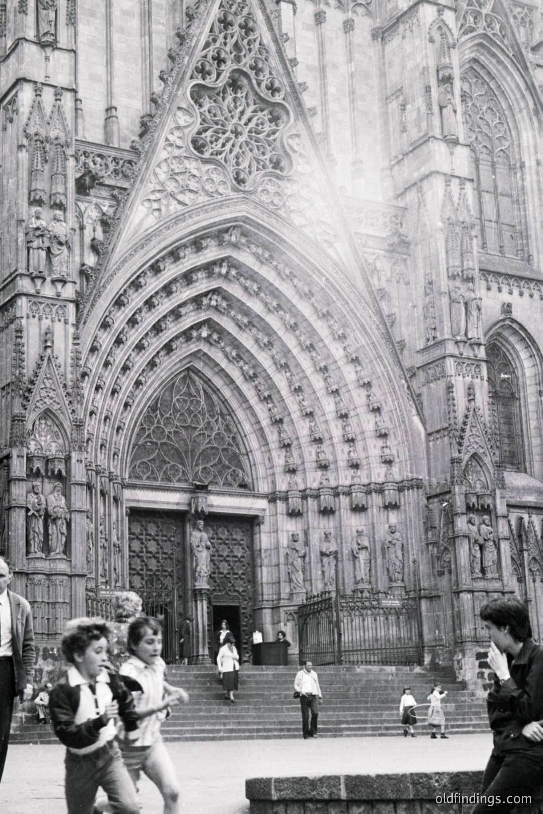 Monumental view of a Gothic cathedral façade, likely Notre Dame, showcasing intricate carvings & a rose window. Several people ascend the wide stone steps; children play near the base. Appears to be a candid street photograph, circa 1970s. High commercial value for design/historical reference.