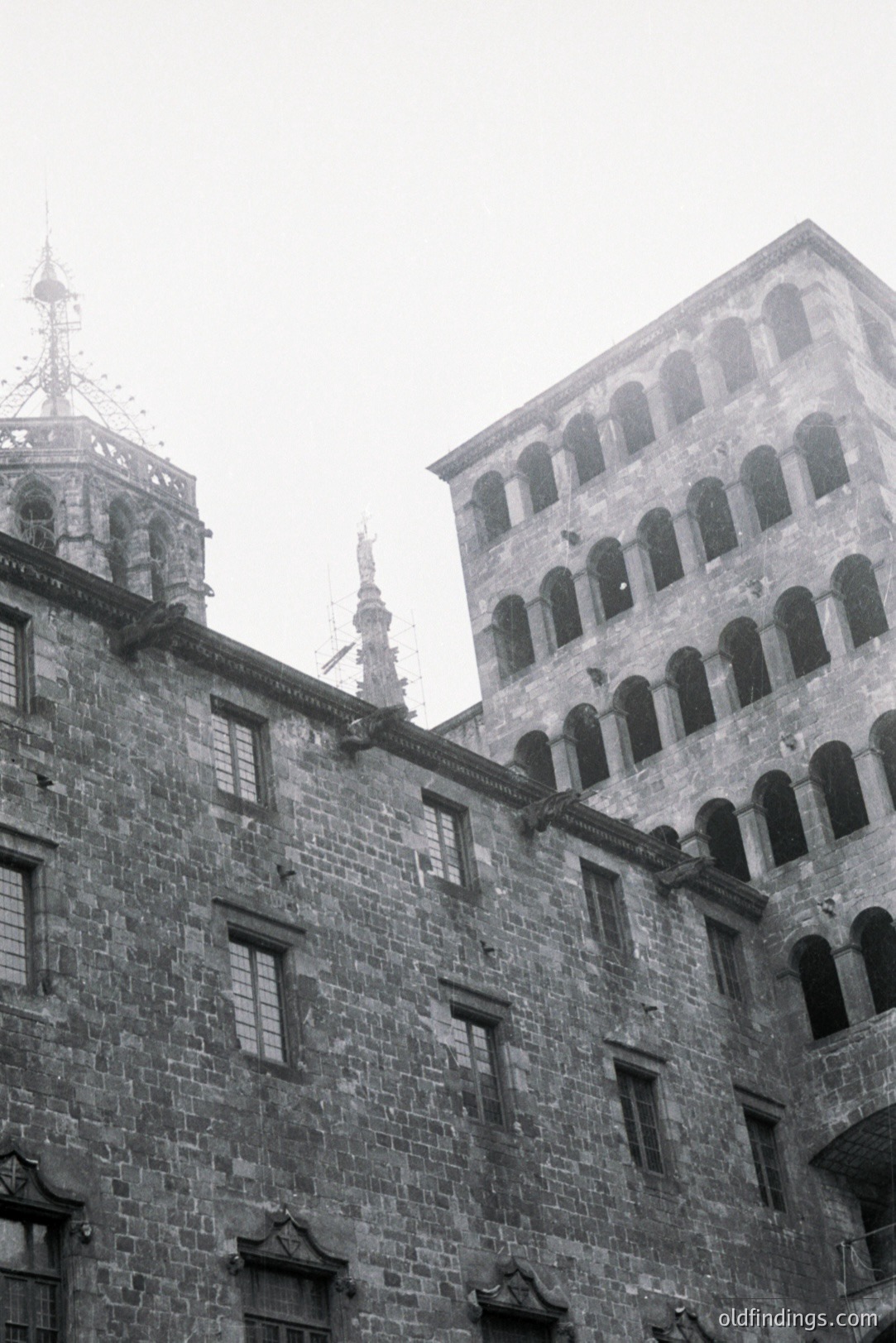 A low-angle, black and white image depicts a portion of a medieval stone building, likely a palace or fortress. Note the tiered arches, intricate stonework, and defensive towers. Architectural details suggest a European origin. Possibly 14th-16th century construction. Aesthetically strong for design or historical context.