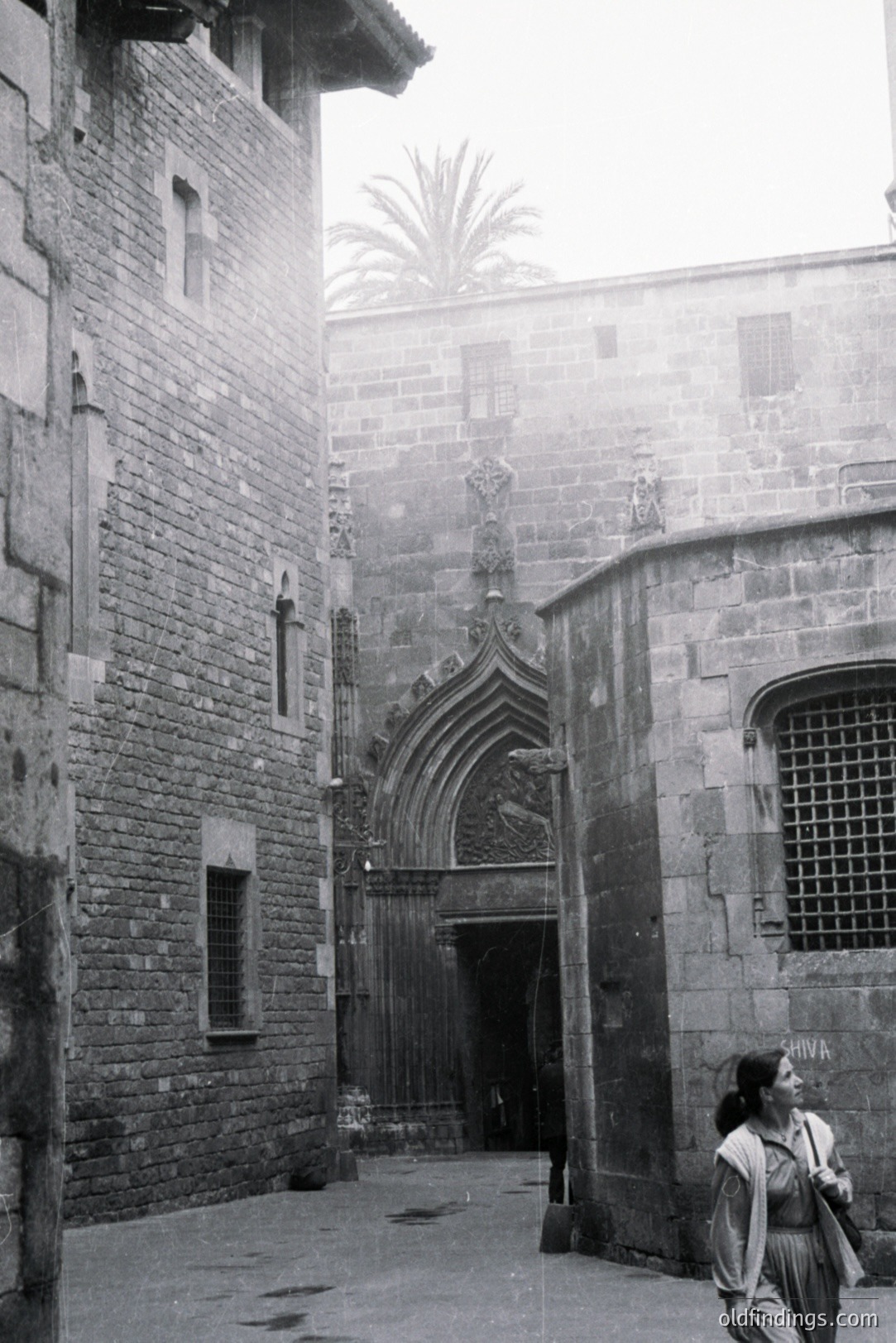 Monochrome image depicts a stone courtyard with ornate Gothic doorway. A lone woman in a long dress walks away, partially obscured by the architecture. Likely European, mid-20th century, potential stock photo for travel or design. Demonstrates textures & light play.