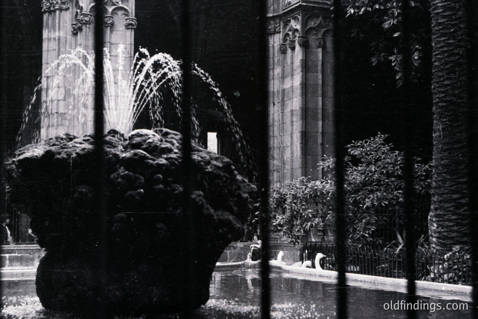 A tiered fountain sits within a courtyard, framed by wrought iron bars. Behind, a Gothic Revival-style building features pointed arch windows. The monochrome image suggests a mid-20th century aesthetic, potentially architectural stock photography. A serene, enclosed space.