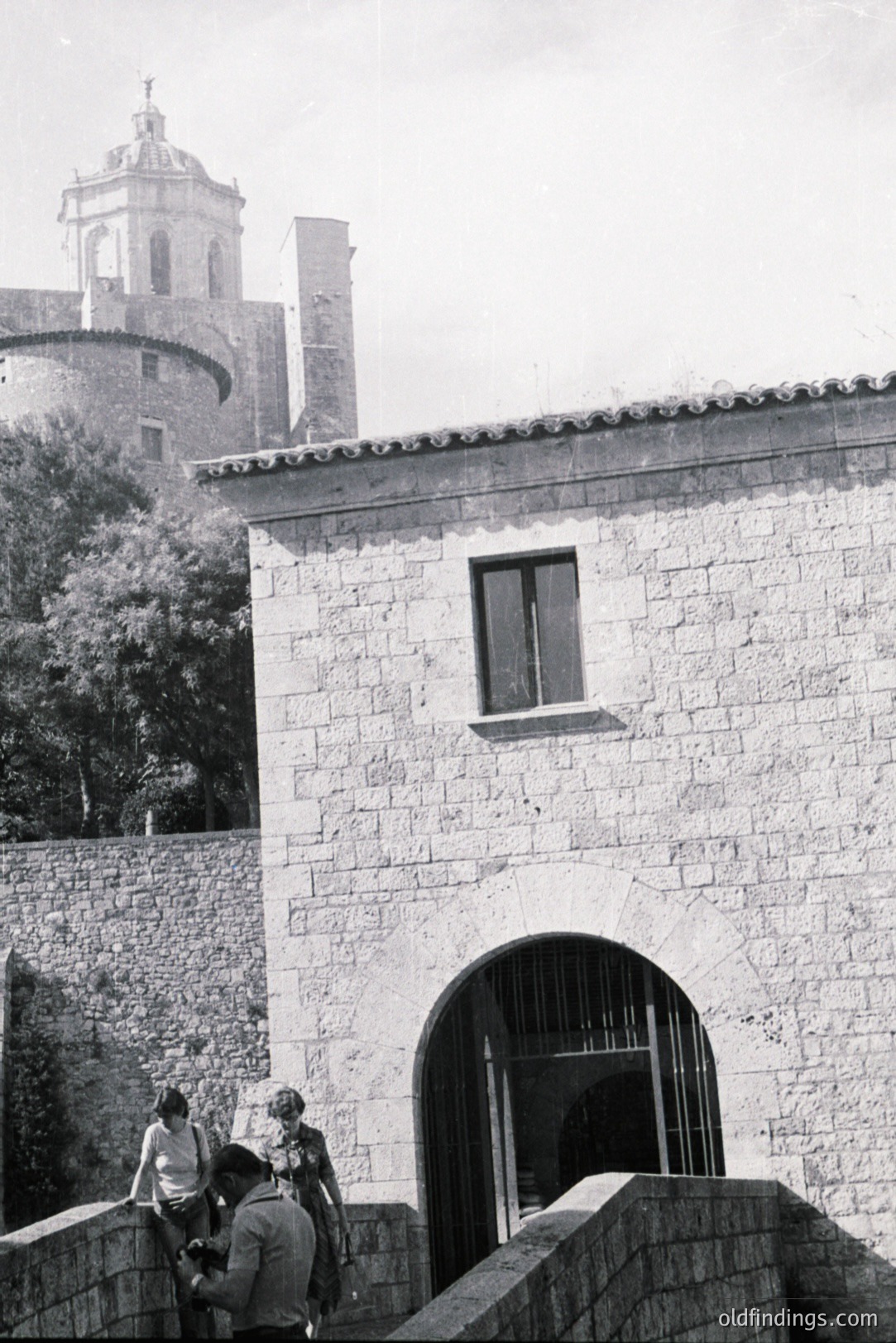 Stone building with arched gateway & partial view of a tower church. Three figures visible – two adults, one child – near the stone wall. Likely a courtyard or fortress entrance. Photographic style suggests the 1960s or 70s, possibly Southern Europe. Interesting architectural detail for design reference.