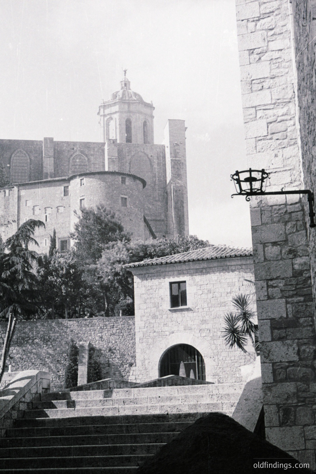 A stone building with an arched opening and single window fronts a tiered landscape featuring a large, historic structure with a central bell tower. Likely Southern Europe, perhaps Italy, circa 1970s, based on photographic style & architecture. Stone steps and foliage enhance perspective.
