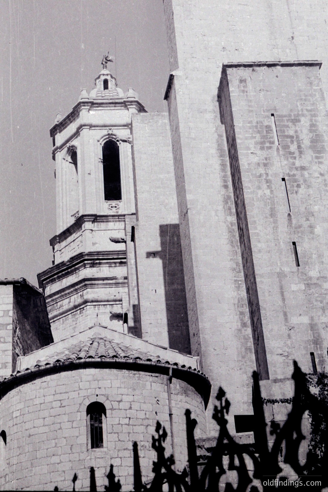 Stone church & bell tower with a tiered design & a cross atop. Arched windows are visible. Likely a European architectural style from the mid-20th century. This image exhibits a strong contrast between light and shadow. Architectural documentation or historical reference material.