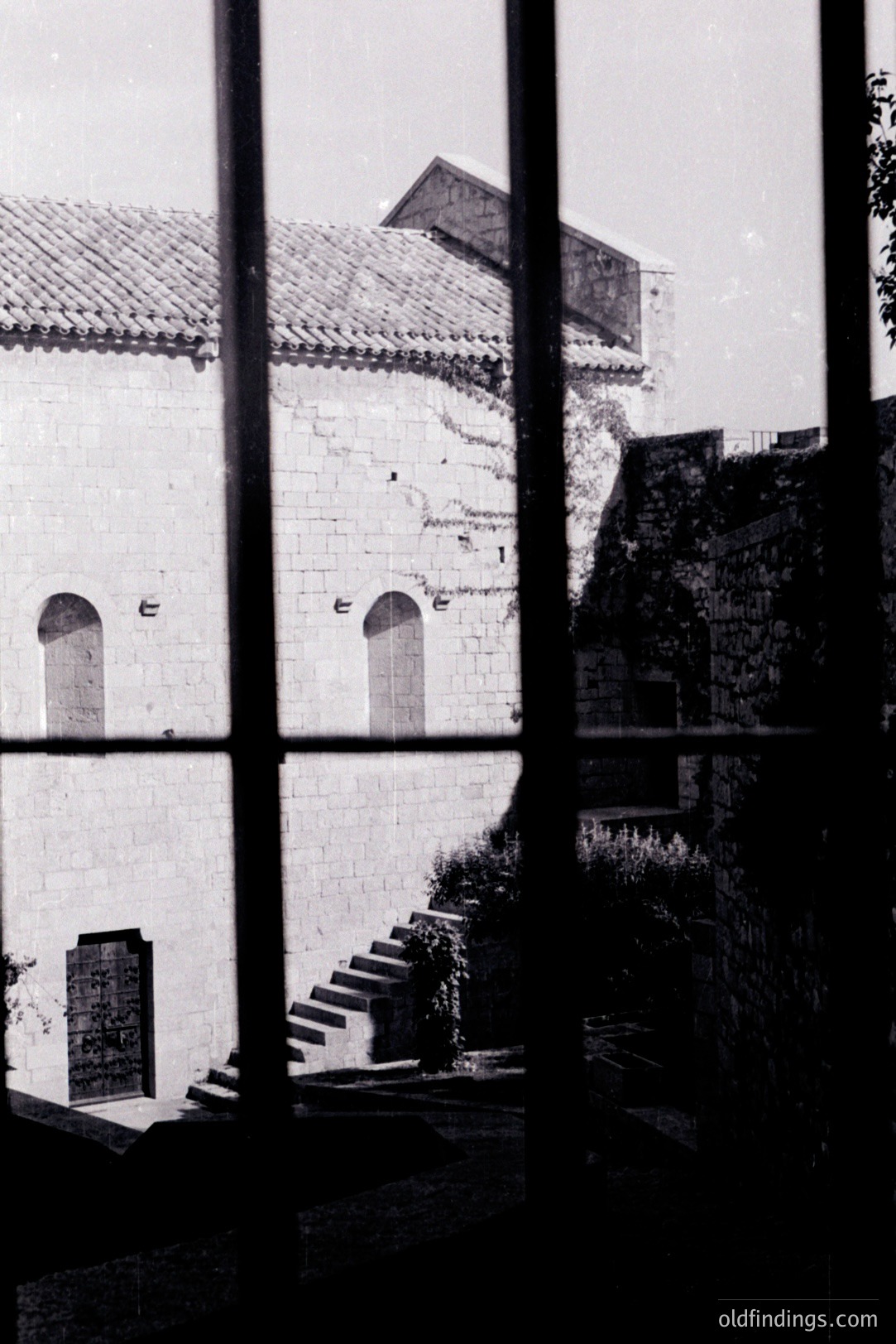 Monochrome image: weathered stone facade with arched windows and staircase framed by window mullions. Ivy climbs the walls, suggesting a historic building, possibly a monastery or villa. Appears to be a European location, likely 20th century. Architectural detail provides design reference.
