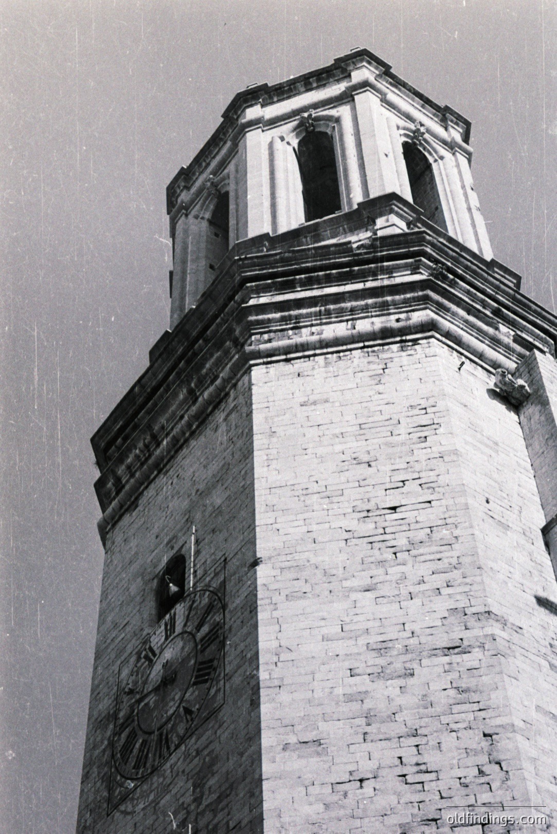 Dramatic low-angle view of a brick tower, featuring a clock face and decorative neoclassical details. The tower's design includes arched windows and a cornice, suggesting a public building or landmark. Likely mid-20th century, possibly architectural study. A vintage aesthetic with visible grain.