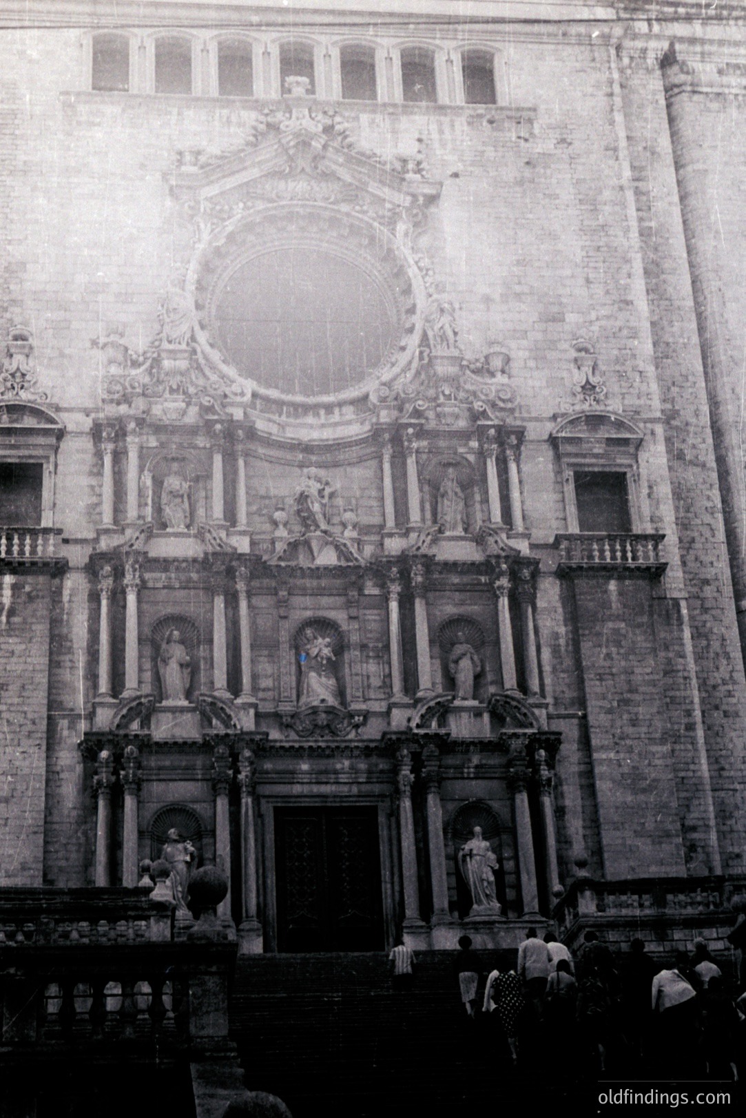 Monumental facade of a baroque-style cathedral features elaborate sculptural detailing, statues, and a central circular window. Crowd of onlookers observed at the base, possibly during a public event. Likely 1960s-70s, film photography aesthetic.