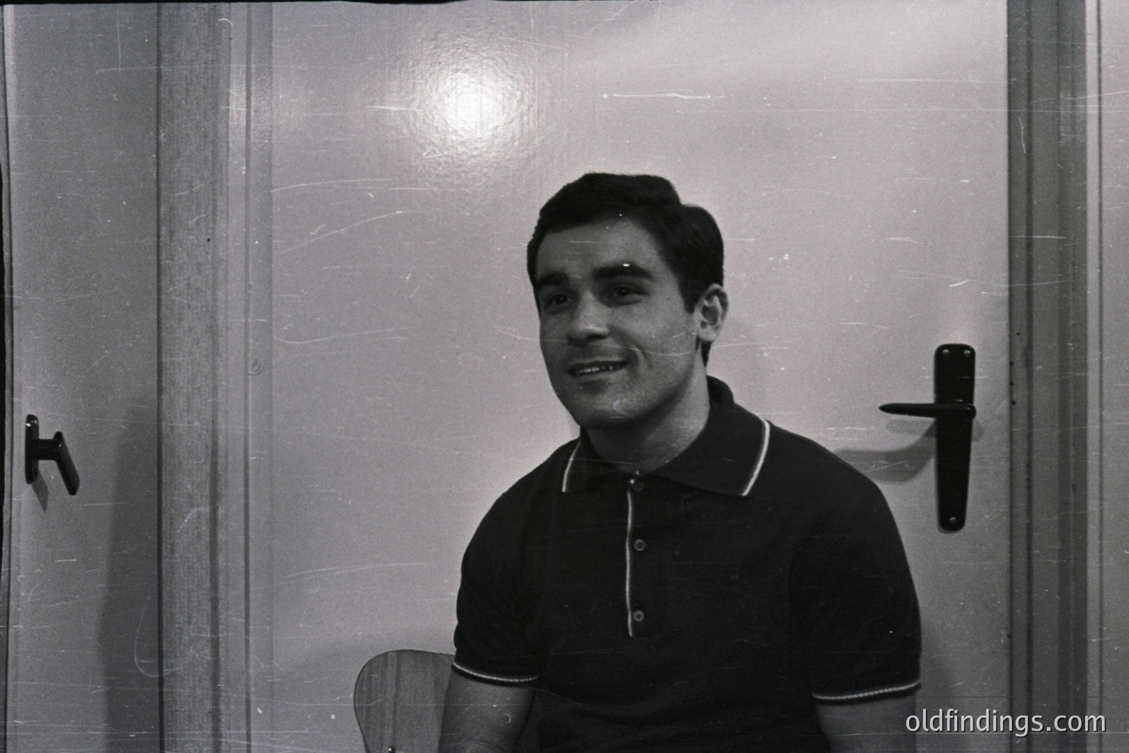 A smiling man in a black polo shirt sits near a door. The photograph’s minimalist setting highlights the subject’s casual style and engaging expression. Likely from the 1960s or 70s, this image offers a glimpse into everyday life. Suitable for archival or design reference.