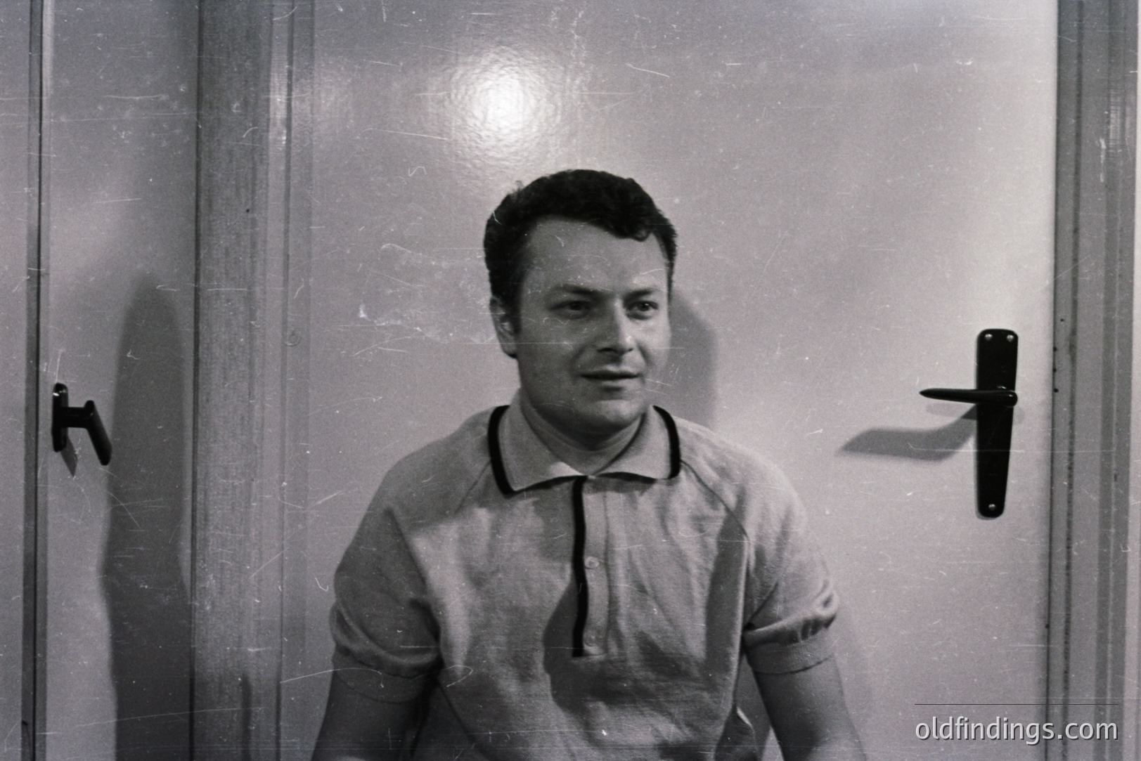 Close-up of a young man in a light-colored polo shirt, posing in front of a plain door. Appears to be a candid, possibly promotional, studio portrait. The door's hardware suggests mid-century design. Minimalist background enhances subject focus. Likely from the 1960s.