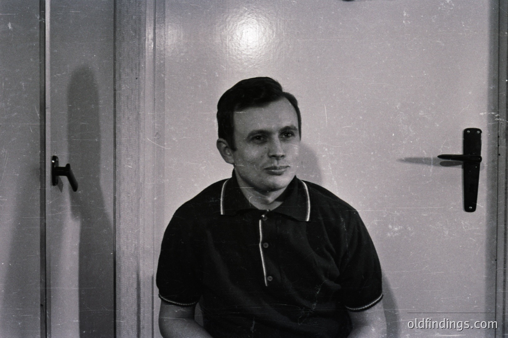 A monochrome portrait of a man seated against a plain wall and door. He wears a black polo shirt with white trim. Simple, geometric hairstyle & neutral expression. Likely mid-20th century portraiture, potentially studio or informal setting. Suitable for design and historical reference.