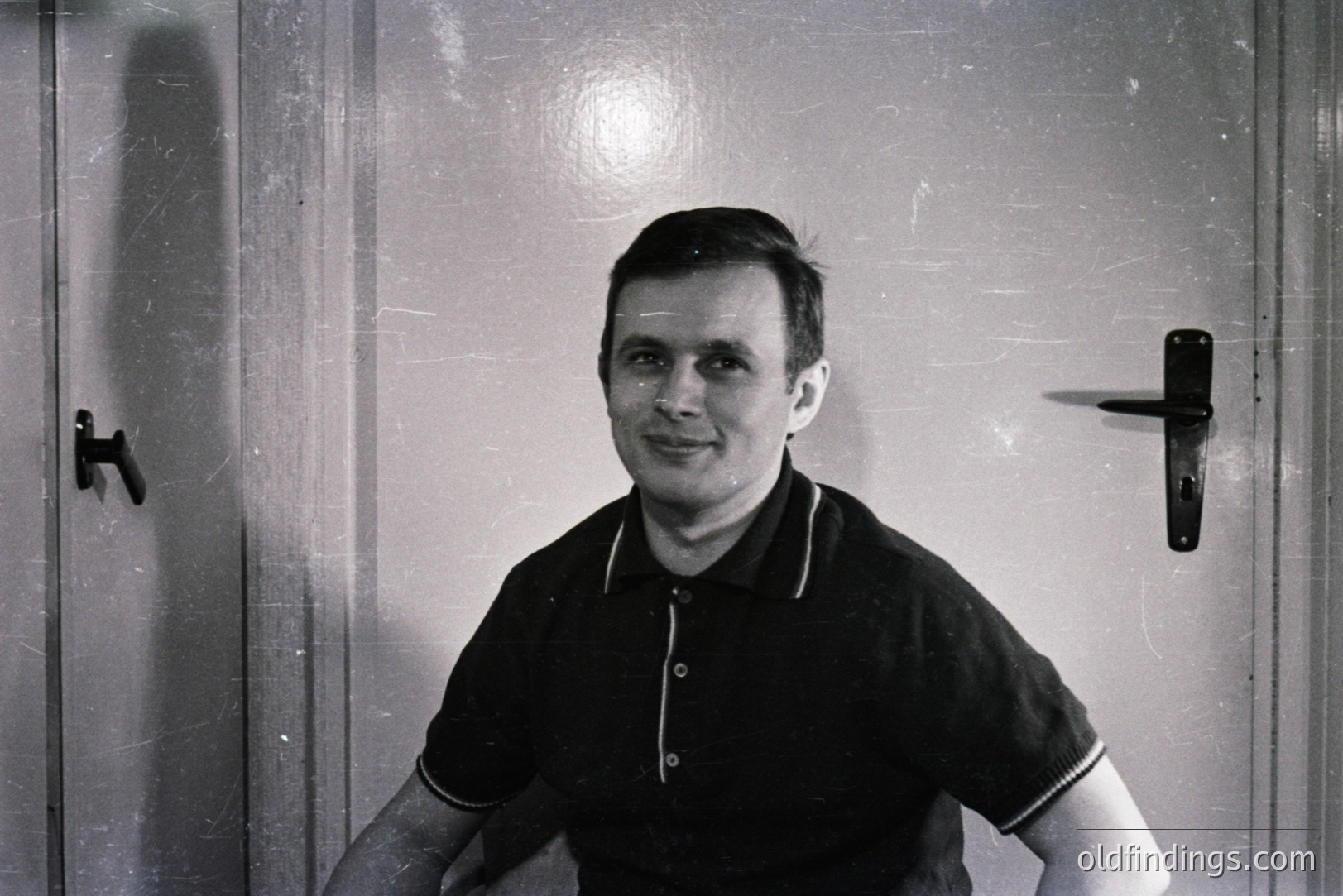 A monochrome portrait of a young man posing in front of a door. He wears a dark polo shirt with a white trim. The door features a prominent, minimalist door handle. Likely taken in the 1960s or 1970s; characteristic of mid-century portraiture. Simple, understated style.