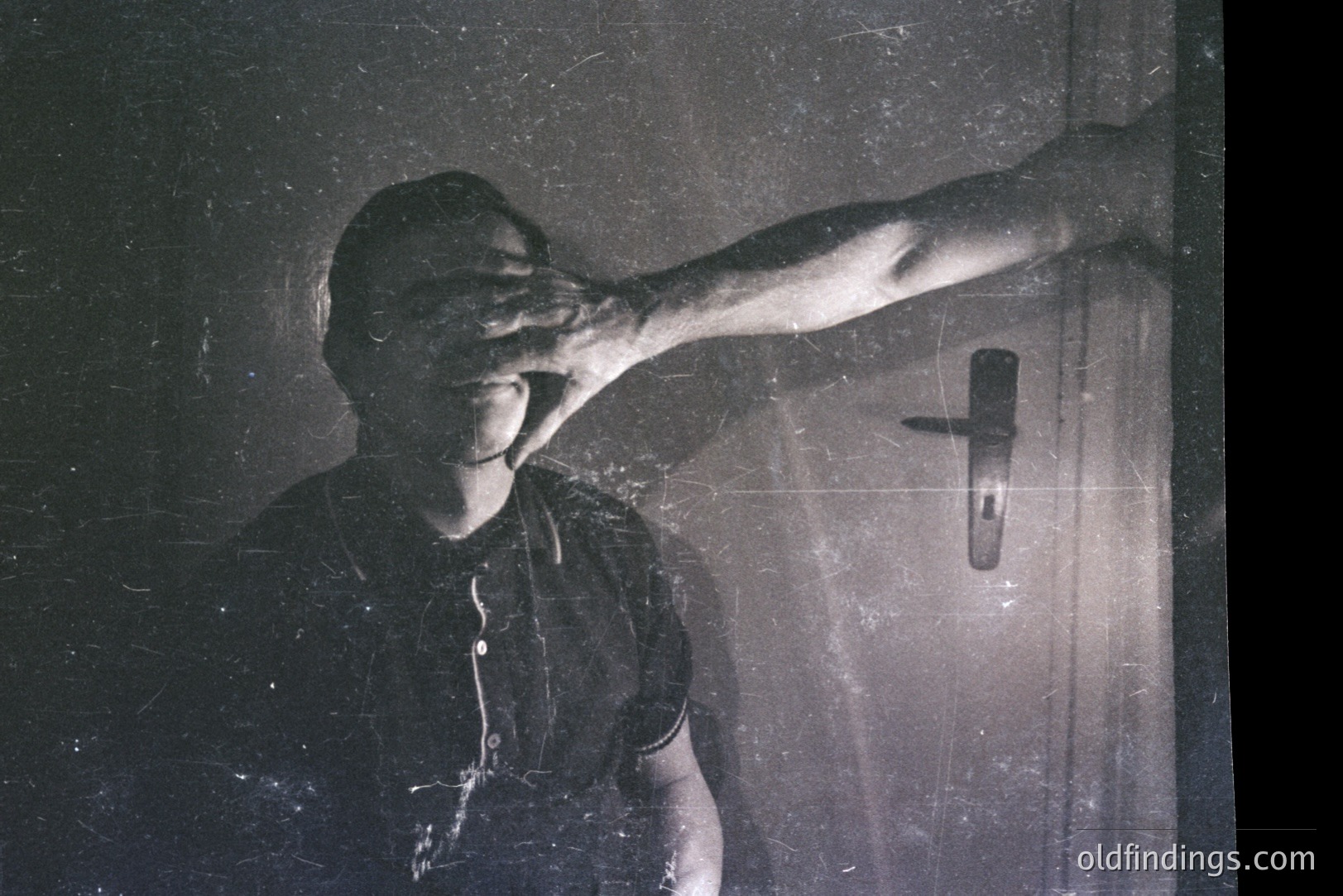 A man blocks his face with his hand in a grainy, high-contrast black and white image. Visible wall and door hardware suggest an interior setting. The subject's clothing indicates a casual style. Image exhibits film grain and dust, possibly indicating age. Likely a candid portrait, potentially 1970s or 80s.