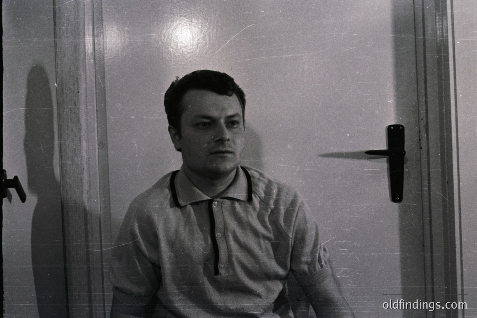 A portrait of a young man in a short-sleeved polo shirt. The photograph, exhibiting film grain and light flares, features a plain wall and door with a prominent door handle behind him. Likely 1960s-70s aesthetic. Suitable for design or historical reference.
