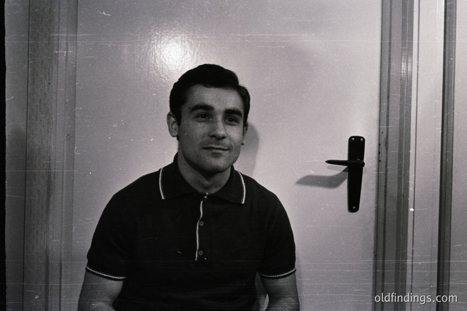 Monochrome portrait of a man in a black polo shirt with white trim. He is positioned against a plain, possibly institutional, wall with a dark metal doorknob visible. Likely a 1960s-70s snapshot. Useful for design, mood boards, or historical context. Simple, direct composition.