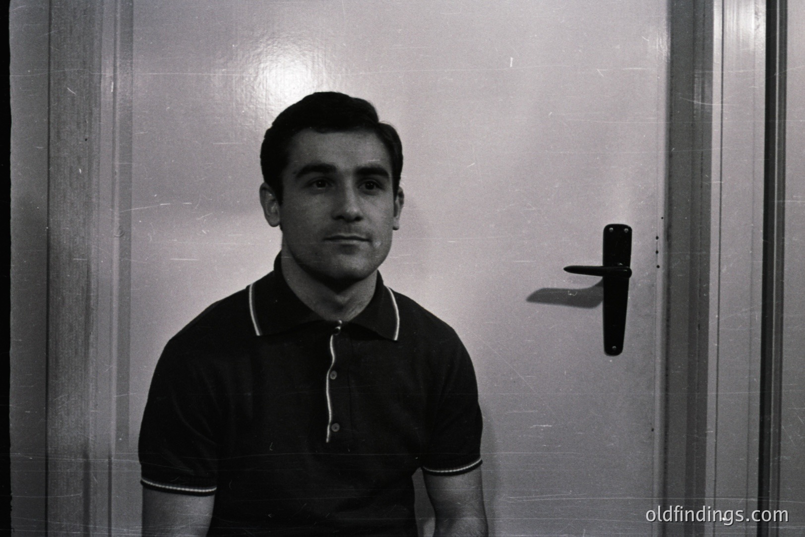 A portrait of a man in a black polo shirt stands against a door. The composition is simple; focus on the subject’s direct gaze. Likely 1960s-1970s style, indicative of a period fashion. Strong potential for design reference & vintage aesthetic appeal. Minimalist and impactful.