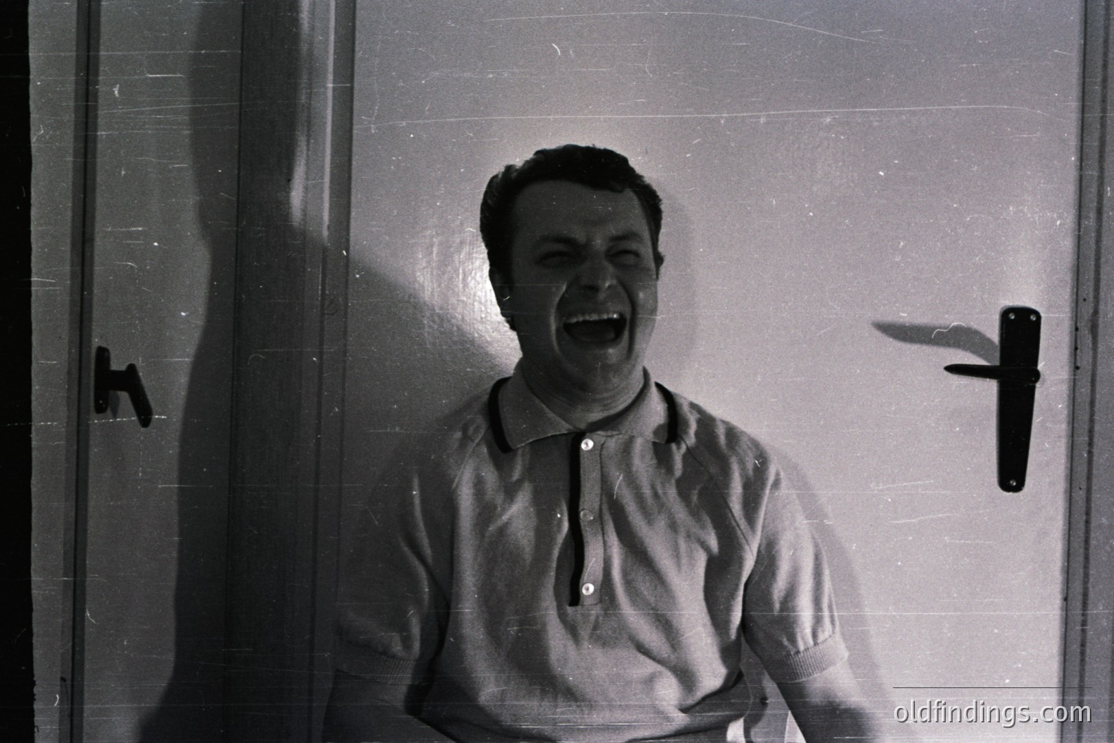 Man with an open mouth, appearing to shout or exclaim. Portrait-style, against a plain door background. He wears a short-sleeved polo shirt. Likely 1960s or 70s. Door hardware suggests a utilitarian design. Potential for stock use showcasing emotion/reaction.