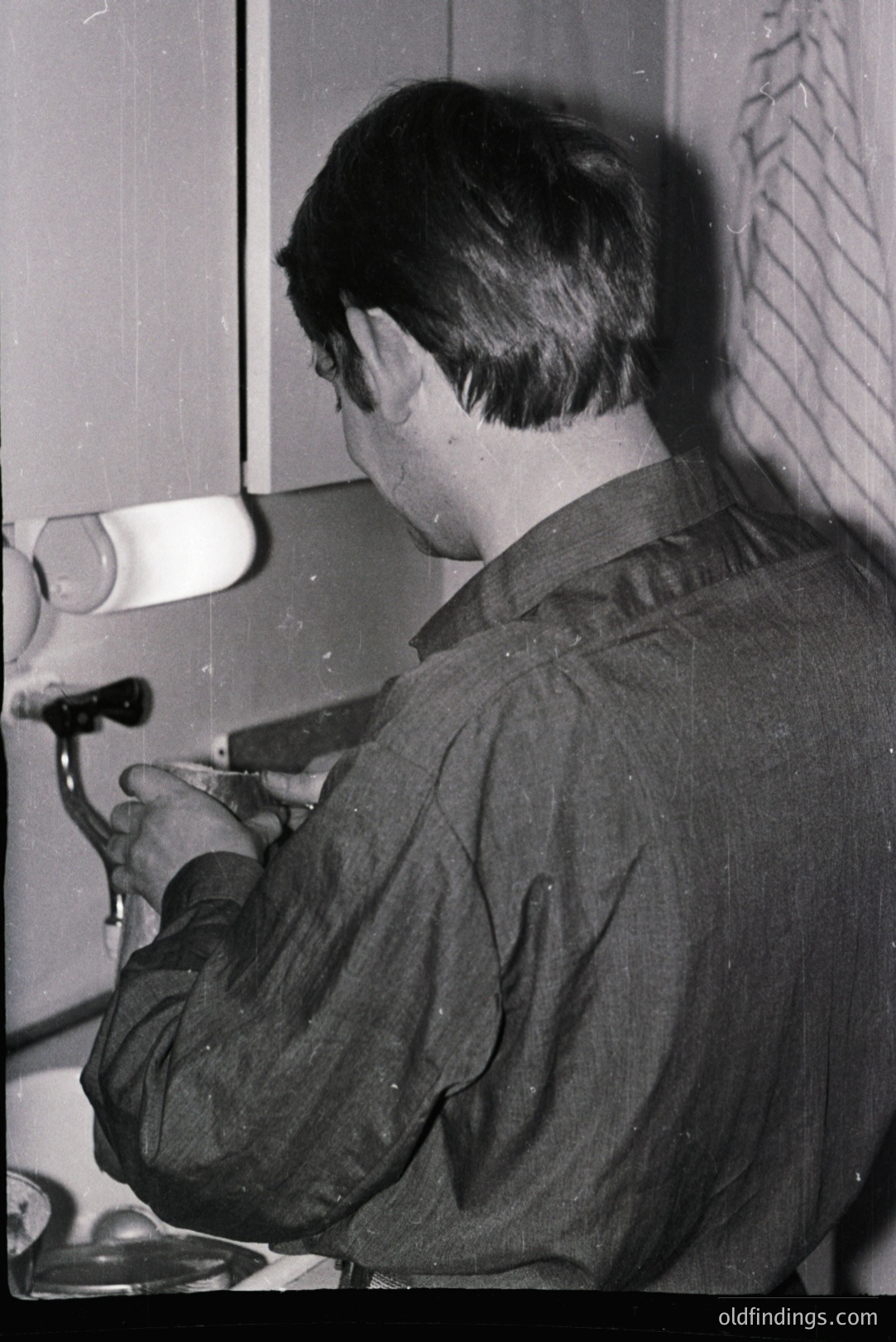 A young man, viewed from behind, meticulously works on a watch or clock mechanism in what appears to be a workshop or repair setting. Tools and a cleaning cloth are visible. The scene evokes a mid-20th century atmosphere. Likely or .