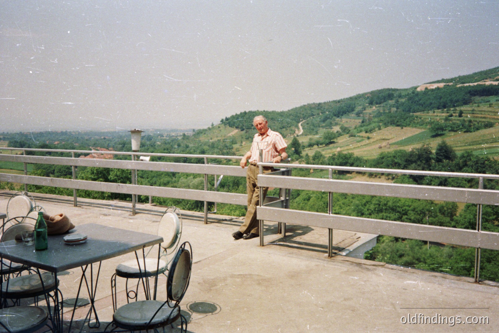 An older man stands near a metal railing overlooking a lush, green, hilly landscape. A table with drinks and cutlery sits nearby on the rooftop terrace. Likely Eastern European, possibly 1970s or 80s aesthetic based on clothing and architecture. A tranquil, panoramic view.