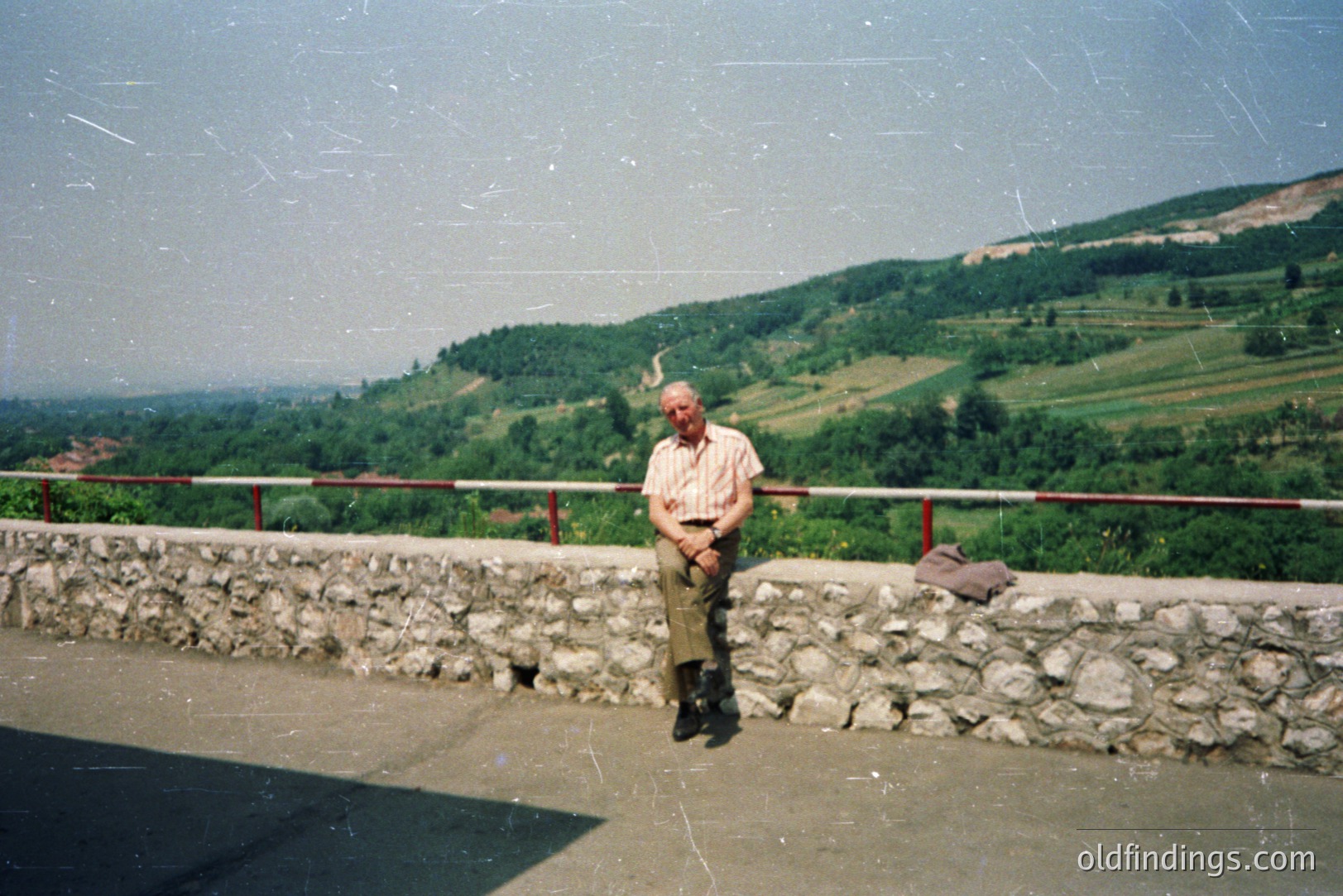 A senior gentleman, wearing a short-sleeved shirt & corduroy trousers, sits on a stone retaining wall overlooking a hillside landscape. The scene features terraced fields and dense vegetation. Likely rural area, possibly Eastern Europe. Appears to be a snapshot from the 1970s or 1980s, evident in film grain.