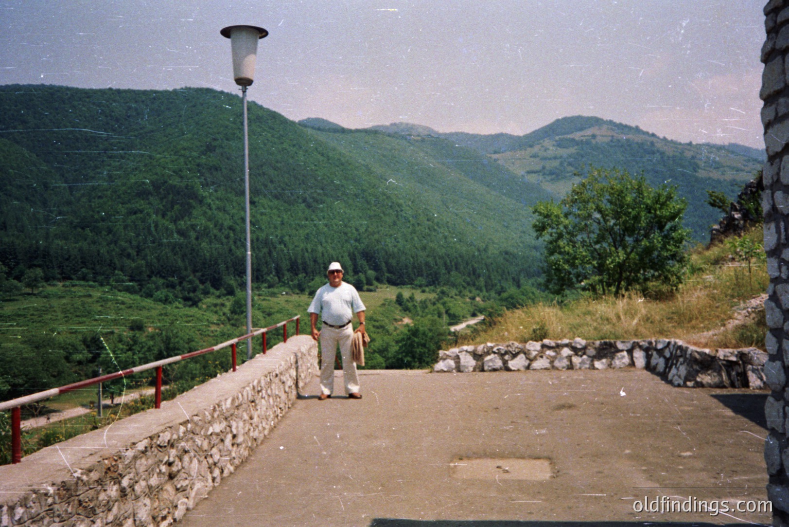 An older man in a white outfit stands on a paved overlook, framed by a stone retaining wall and a metal lamppost. Verdant, forested hills extend into the background, suggesting a mountainous landscape. Appears to be a roadside vista, possibly in the Balkans. Likely 1970s or 80s film.