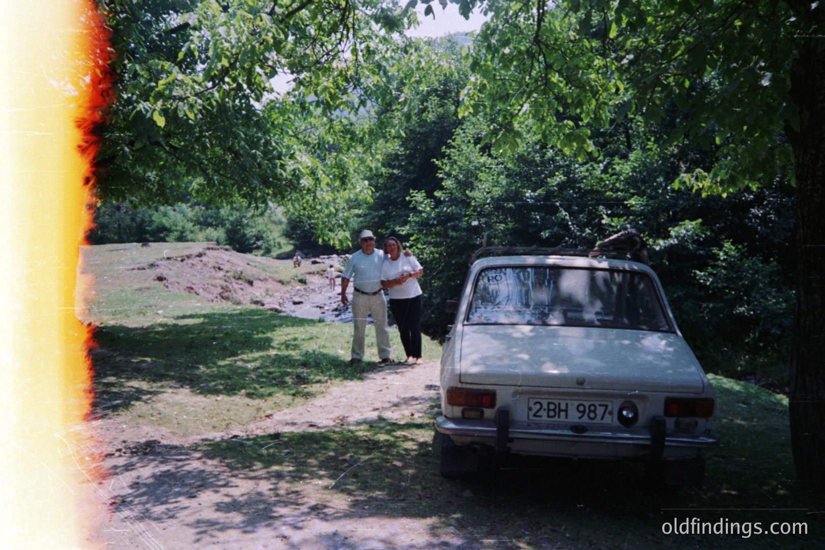 A vintage sedan, likely a Lada, sits on a dirt road flanked by dense woodland. Two figures, a man in a hat and a woman, stand nearby. The plate reads "2-BH-987," suggesting Bulgaria. The scene evokes a 1970s or 80s holiday atmosphere. Photo's border displays light leakage.