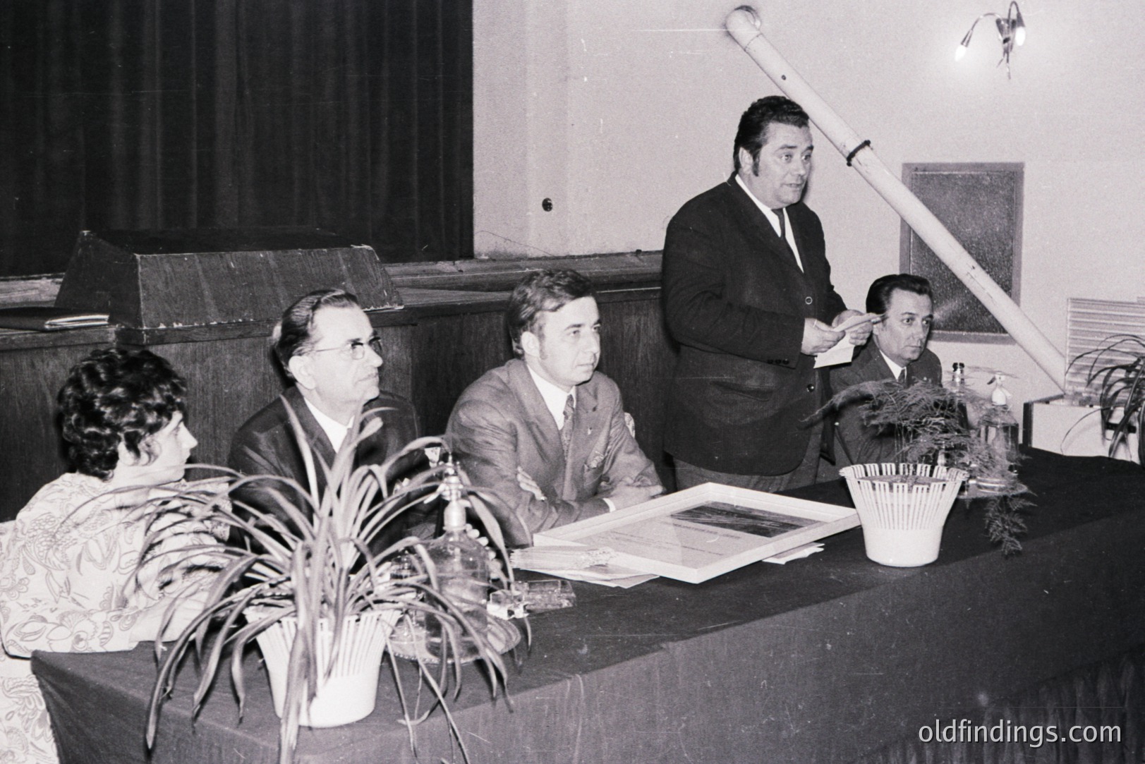 A formal gathering features four people seated at a table. A man stands speaking, likely presenting to the seated group. The setting appears to be a hall or auditorium, evident from the stage backdrop. Likely 1960s-1970s, judging by the attire and photographic style.